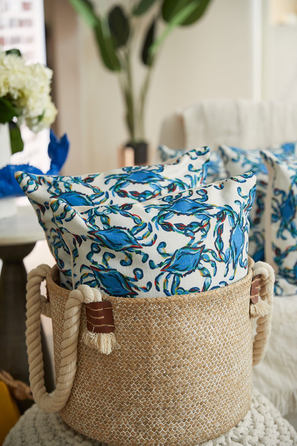 A person in a light blue shirt and beige pants sits, hugging the Crab Craze Shades of Blue Lumbar Pillow, featuring a colorful blue and green leafy pattern. The shot is partially cropped to reveal only their mouth and hands.