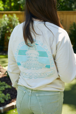 A person with long brown hair stands outside in light green pants and a Charleston Pineapple Crewneck Sweatshirt, which features a graphic of the iconic fountain and "Charleston, South Carolina" printed on the back.