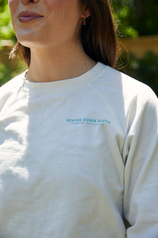 A woman stands outdoors in sunlight, wearing a light-colored Charleston Pineapple Crewneck Sweatshirt with “SEWING DOWN SOUTH CHARLESTON, SOUTH CAROLINA” embroidered on the chest.