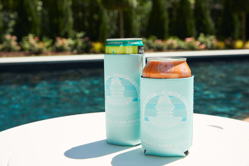 A man in a white shirt and teal shorts sits by a pool. In the foreground, a can with an orange top is in a CHS Pineapple Koozie (4 PACK) on a white table, with another empty drink cooler nearby.