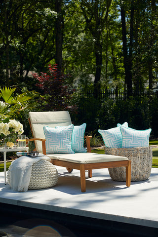 A wooden outdoor lounge chair with a Mint Greek Key Outdoor Pillow sits on a patio next to a basket, pouf with blanket, and side table holding flowers, a drink, and sunglasses amid lush greenery.