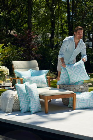 A man in a light blue shirt and white shorts arranges the Mint Greek Key Outdoor Pillow, made with SDS Shield® Outdoor Fabric, on a wooden chair and ottoman by the pool as sunlight filters through nearby trees and greenery.