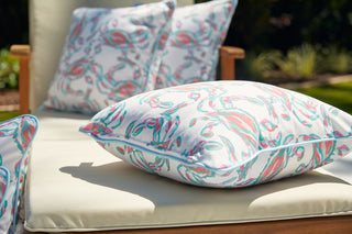 A cozy lounge chair with white cushions features the Coral Crab Craze Outdoor Pillow by Sewing Down South™, accentuated by vibrant pink, teal, and green brushstrokes. The durable SDS Shield® Outdoor Fabric complements the sunny garden setting.