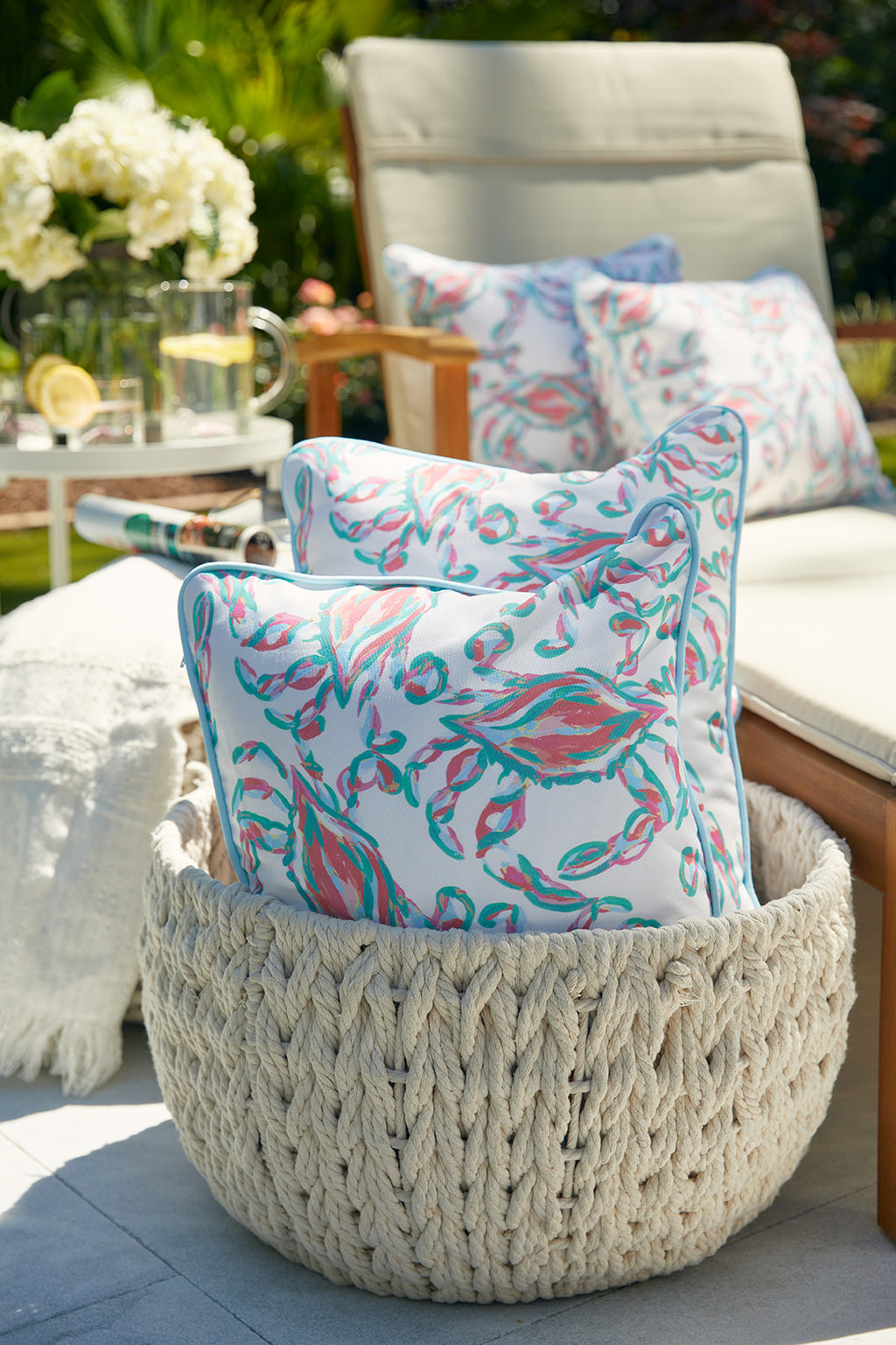 A patio setting includes a wicker basket with colorful floral pillows made from SDS Shield® Outdoor Fabric and additional cushions on a wooden lounge chair, featuring a Coral Crab Craze Lumbar Outdoor Pillow. A glass pitcher with glasses rests on a small table surrounded by flowers.
