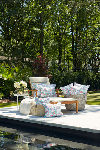 The outdoor scene showcases a plush lounge chair and ottoman with the Coral Crab Craze Lumbar Outdoor Pillow. A small table with white flowers complements the lush greenery, trees, and pool edge in the foreground.