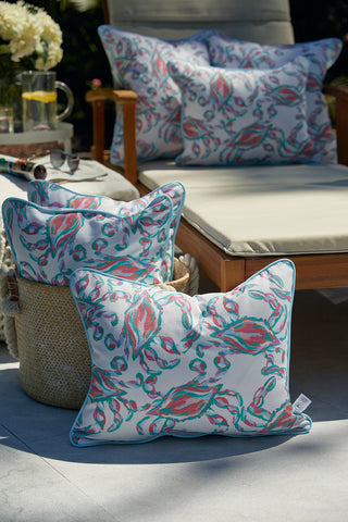 A patio setting includes a wicker basket with colorful floral pillows made from SDS Shield® Outdoor Fabric and additional cushions on a wooden lounge chair, featuring a Coral Crab Craze Lumbar Outdoor Pillow. A glass pitcher with glasses rests on a small table surrounded by flowers.