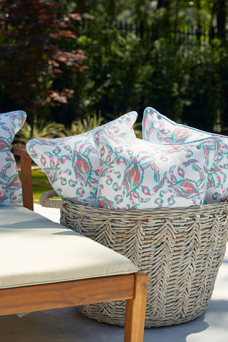 A wicker basket holds Sewing Down South™ cushions in pastel colors beside a cushioned wooden chair on the patio. Sunlight filters through the trees, creating a serene ambiance and highlighting the beauty of Coral Crab Craze Outdoor Pillows.