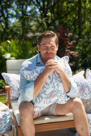 A man in a light blue shirt and beige shorts smiles outdoors, holding a Coral Crab Craze Outdoor Pillow featuring a leaf pattern. Around him are Sewing Down South™ design pillows made from SDS Shield® Outdoor Fabric, set against lush greenery.