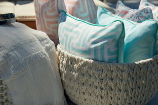 A woven basket holds decorative pillows, such as the Mint Greek Key Outdoor Pillow, and a cream textured throw, creating a cozy outdoor seating area with pale geometric patterns and aqua accents.