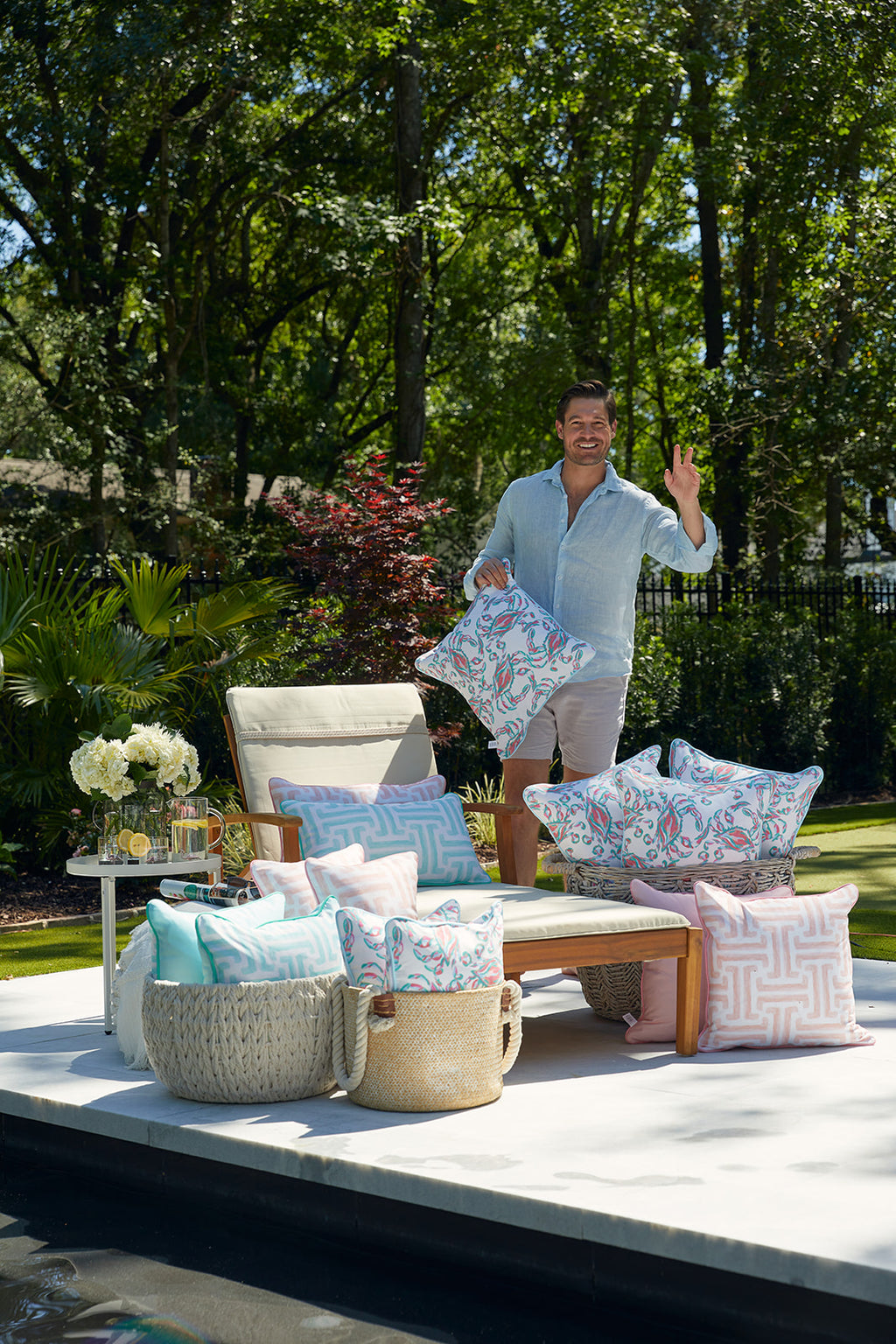 The outdoor patio chair, adorned with two colorful pattern pillows like the Coral Crab Craze Outdoor Pillow, showcases a vibrant mix of pink, blue, and green hues. Surrounded by lush greenery and sunlit leaves, it crafts a peaceful and welcoming ambiance.