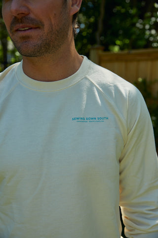 A man wearing a light-colored Charleston Pineapple Crewneck Sweatshirt with "SEWING DOWN SOUTH" and smaller text on the chest stands outdoors in sunlight, surrounded by greenery and a wooden fence.