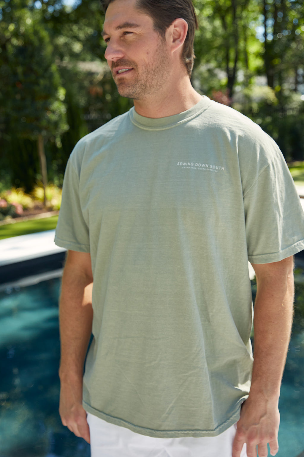 By a pool surrounded by lush greenery, someone wears the comfy WWWMS Short Sleeve Tee in Sandstone, featuring a unisex fit made of 100% ring-spun cotton. Bold white letters on the back question, WHATS WRONG WITH MY SEWING?.
