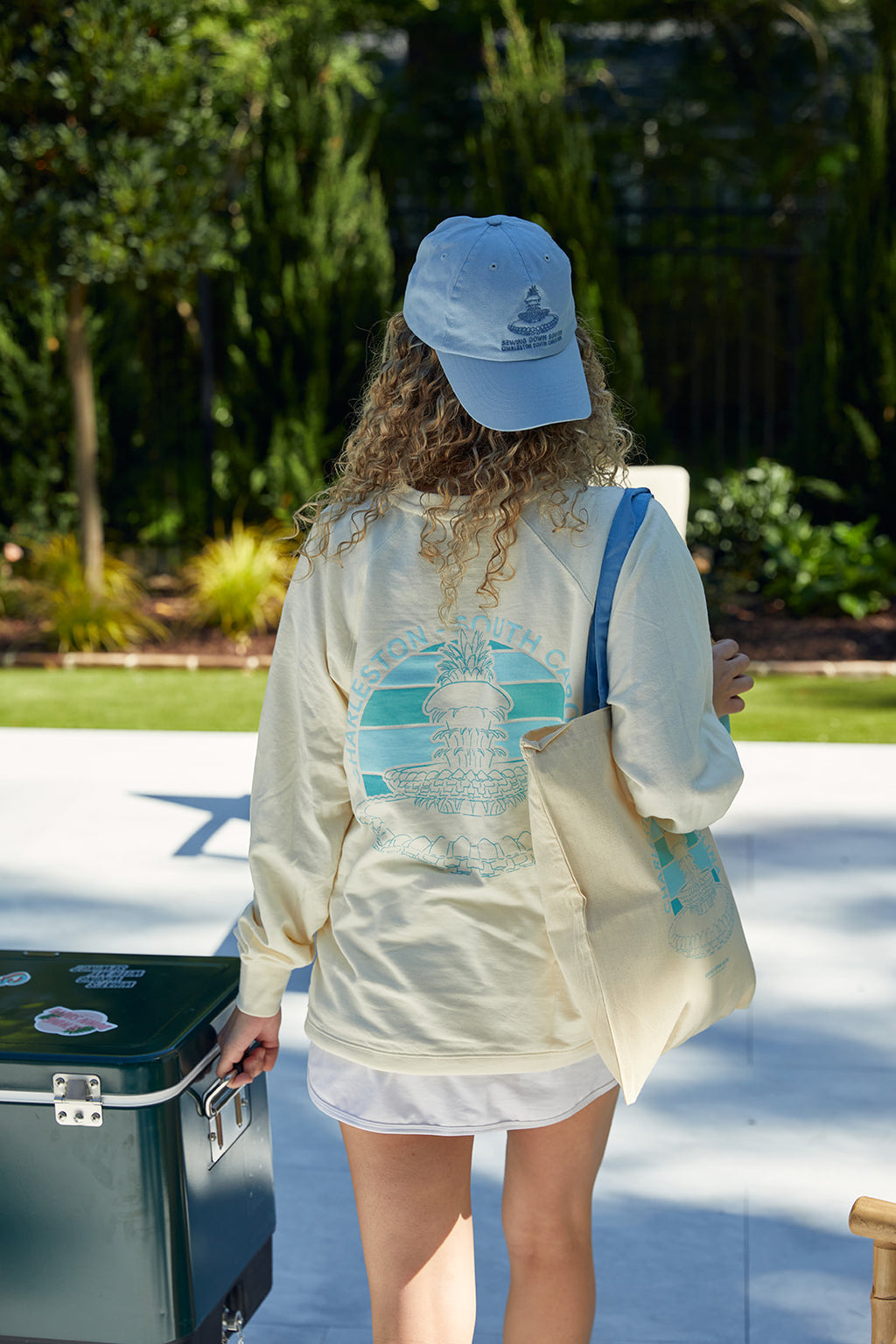 A person with curly hair in a light shirt and cap strolls through a vibrant garden, carrying the Charleston Pineapple Tote. Made from natural cotton with reinforced stitching, it features a lighthouse design while they hold a green container amidst lush greenery.