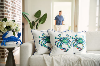 A cozy living room features the Coastal Collection with a white sofa adorned by The King Crab Pillow with Navy. A blue crab sculpture sits on a side table next to white flowers, while someone wearing a navy shirt stands in the background, enhancing the serene coastal vibe.