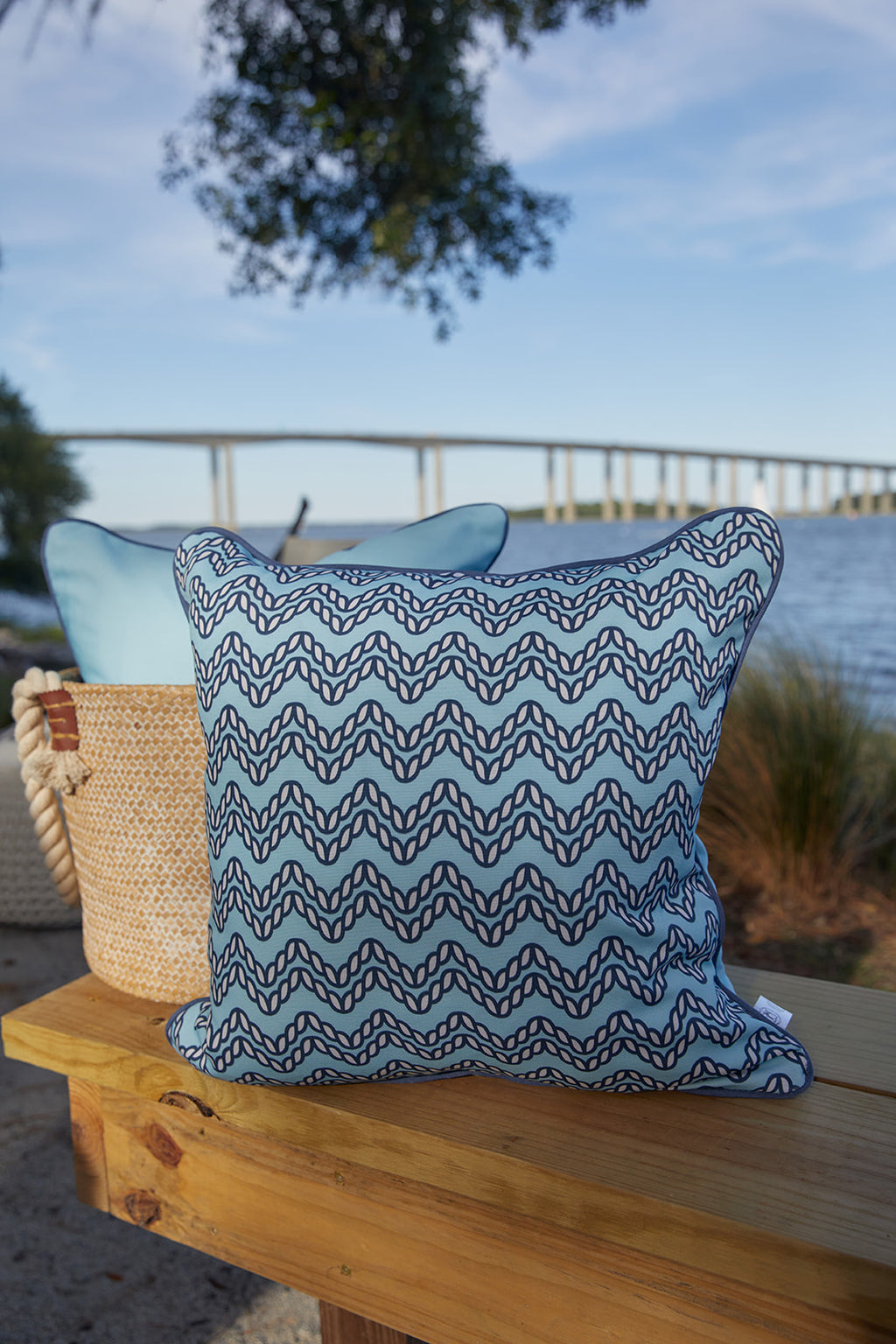 A wooden chair with a Dockside Chevron Ropes Outdoor Pillow sits by the water’s edge, with a bridge in the background and a blanket and basket nearby, creating a relaxed outdoor setting.