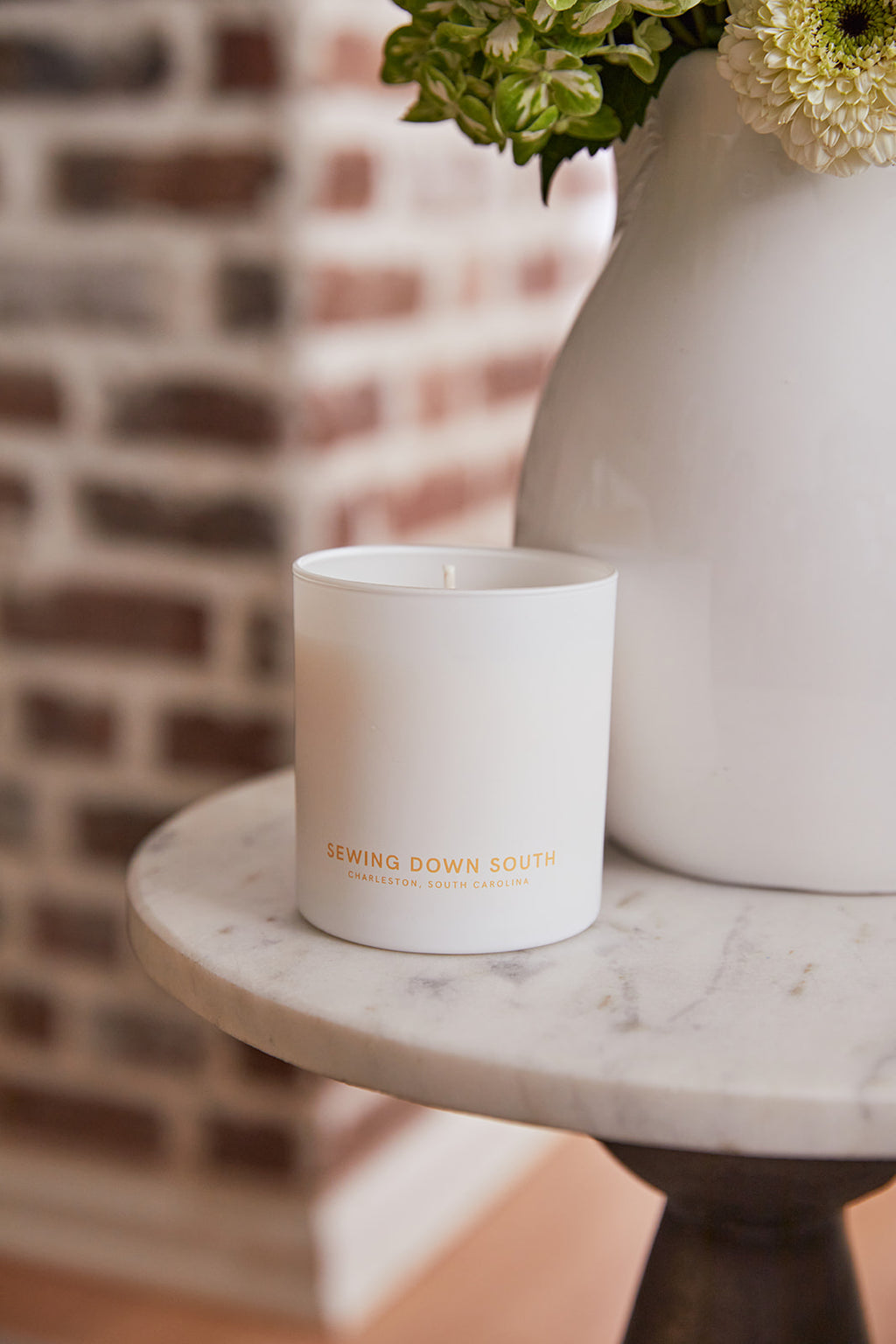 A man in a light blue button-up shirt smiles, holding a white cup towards the camera. The warmly lit room features a lamp with a red base, casting a soft glow on abstract black curtain shapes, reminiscent of the sweet vanilla and buttercream aura from No. 5 - Sugar Cookie Candle.