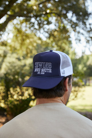 Outdoors, a person in a beige shirt and an SDS Collegiate Trucker hat (Navy and White) with an embroidered SDS logo faces away. Sewing Down South, Est. 2018 is boldly displayed on the cap, surrounded by leafy trees and sunlight.