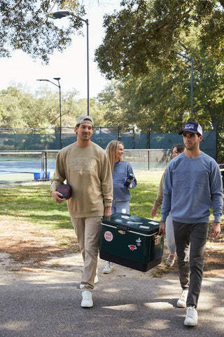 On a sunny day, a group walks outdoors by a tennis court. Two men in casual sweatshirts and pants are in the foreground; one carries a cooler and holds a football, while the other wears an SDS Collegiate Trucker hat in navy and white. Trees can be seen in the background.