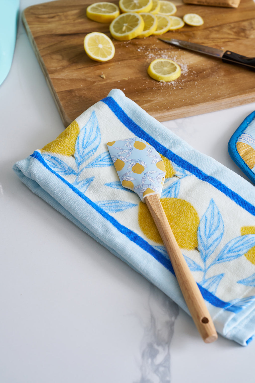 A man in a blue patterned shirt stands in a bright kitchen, a Lemons in a Line Kitchen Towel (2 pack) draped over his shoulder. He holds a lemon near the marble counter with cookies, sunlight streaming through the glass door to the deck.