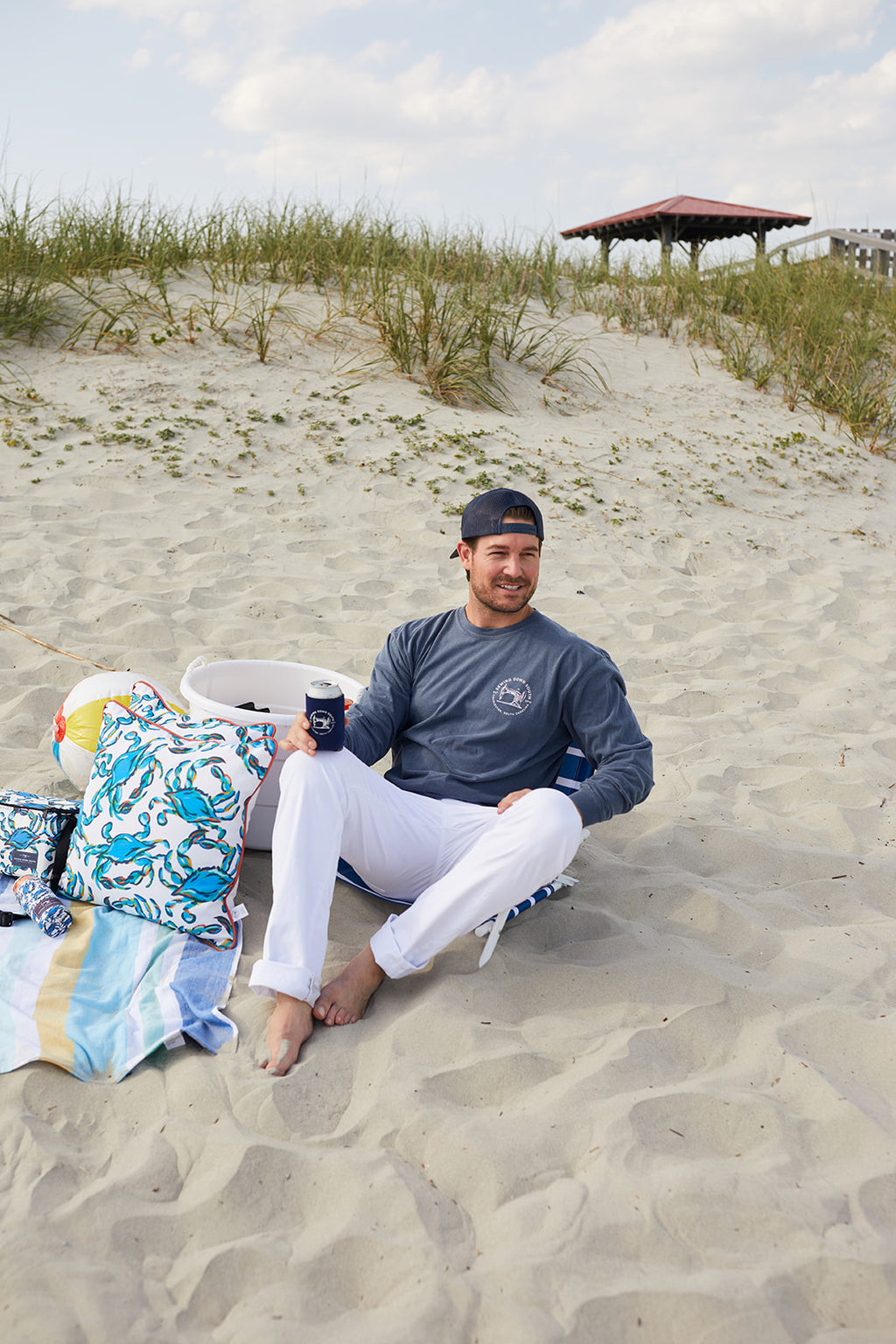 Strolling the beach with a drink and beige tote, a person sports the SDS Vintage Logo Long-Sleeve Tee in Denim and a blue cap. The ocean backdrop highlights this unisex garment-dyed cotton shirt from Sewing Down South.