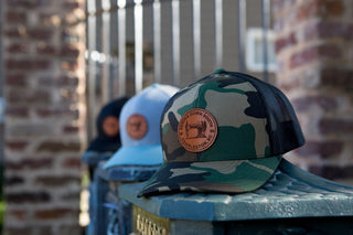The Sewing Down South Leather Patch Trucker in camo, featuring a trucker snapback with vintage logo appeal, is displayed on a stone ledge. Sunlight casts shadows on the blurred metal gate and brick pillar in the background, while white and black hats linger nearby.