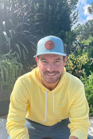 A man in a yellow hoodie smiles, wearing a blue cap with an adjustable snapback while sitting outdoors surrounded by lush green plants. Under the clear blue sky, his hat subtly showcases camo print: the Sewing Down South Leather Patch Trucker, Blue + Black Mesh.