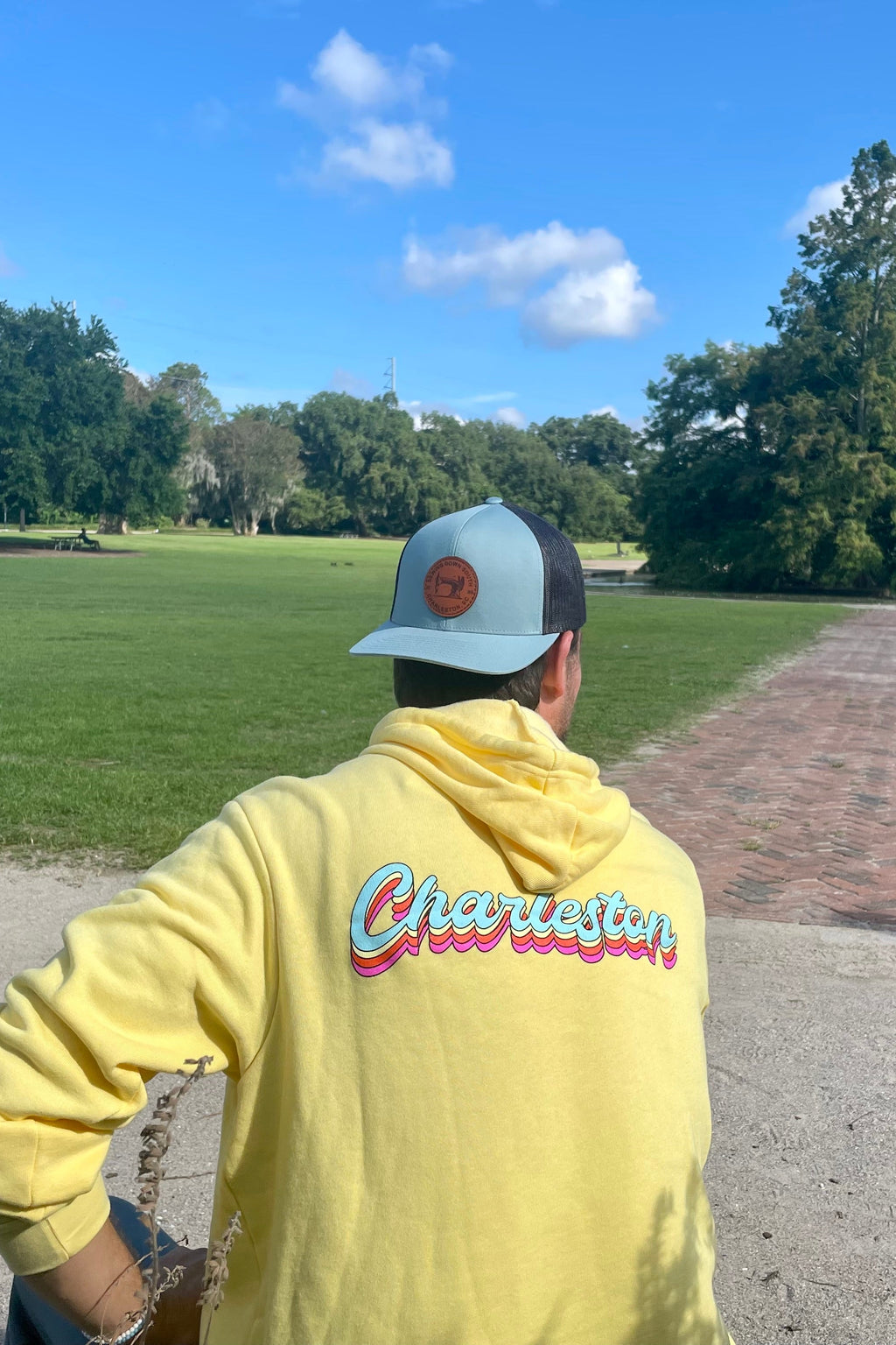 A man in a yellow hoodie smiles, wearing a blue cap with an adjustable snapback while sitting outdoors surrounded by lush green plants. Under the clear blue sky, his hat subtly showcases camo print: the Sewing Down South Leather Patch Trucker, Blue + Black Mesh.