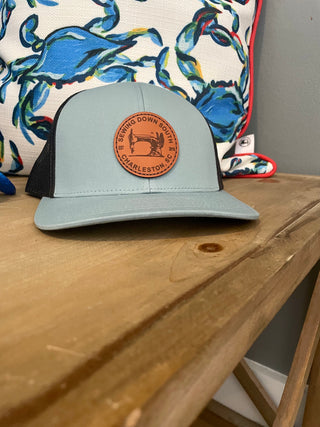 A light gray snapback, the Sewing Down South Leather Patch Trucker with a blue logo and black mesh, rests on a wooden surface. Behind it is a pillow featuring a colorful crab design.