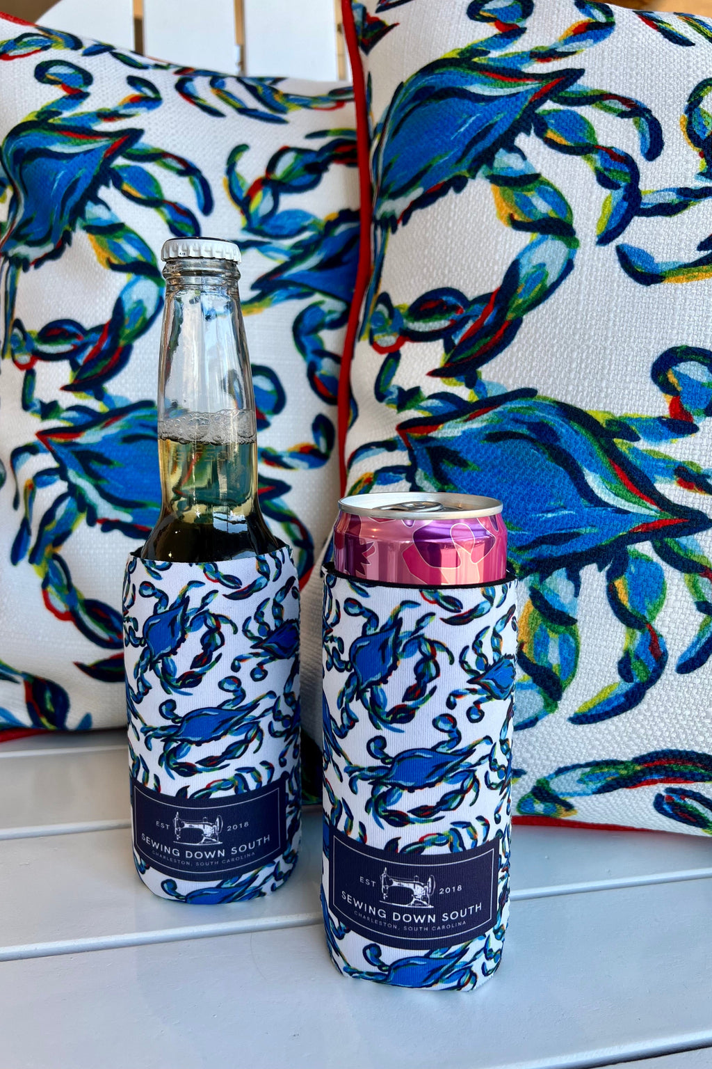 A can in a Crab Craze Koozie, 4 PACK (Skinny or Standard), featuring colorful abstract designs, rests on a blue and white towel inside a white tote bag in a bicycle basket, with part of the bike and brick sidewalk visible.