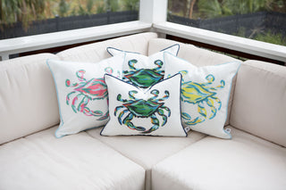 Three Outdoor King Crab Lumbar Pillows with Navy, made from SDS Shield® Outdoor Fabric, feature colorful crab illustrations in pink, green, and yellow. They are placed on a light-colored outdoor sofa surrounded by greenery on a patio.