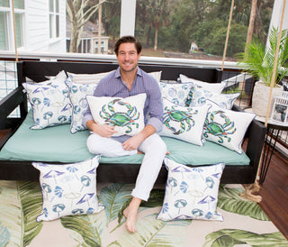 A person sits barefoot on a light blue daybed on a porch, surrounded by Sewing Down South™ Outdoor King Crab Lumbar Pillow with Navy. Trees and greenery create a serene backdrop, reflecting off the durable SDS Shield® Outdoor Fabric of the cushions.