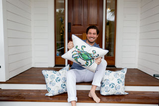 A person on porch steps holds an Outdoor King Crab Lumbar Pillow with Navy, crafted from SDS Shield® Outdoor Fabric. Nearby are two circular-patterned outdoor pillows, featuring the Sewing Down South™ design, set against a wooden door and white siding with plants in the background.