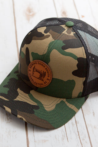 The Sewing Down South Leather Patch Trucker in camo, featuring a black mesh back and a brown circular patch with a vintage logo, rests on a white wooden surface.