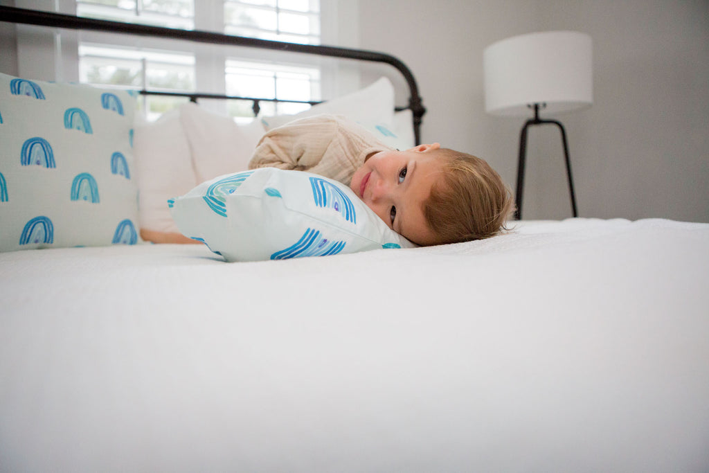 On a bed, Toddler Sleeping Pillow Cases display a front white pillow, a blue and white striped one in the middle, and three colorful ones featuring blue rainbows, yellow school buses, and pink rainbows. These organic, machine washable pillows are complemented by a thoughtful card on the bed.