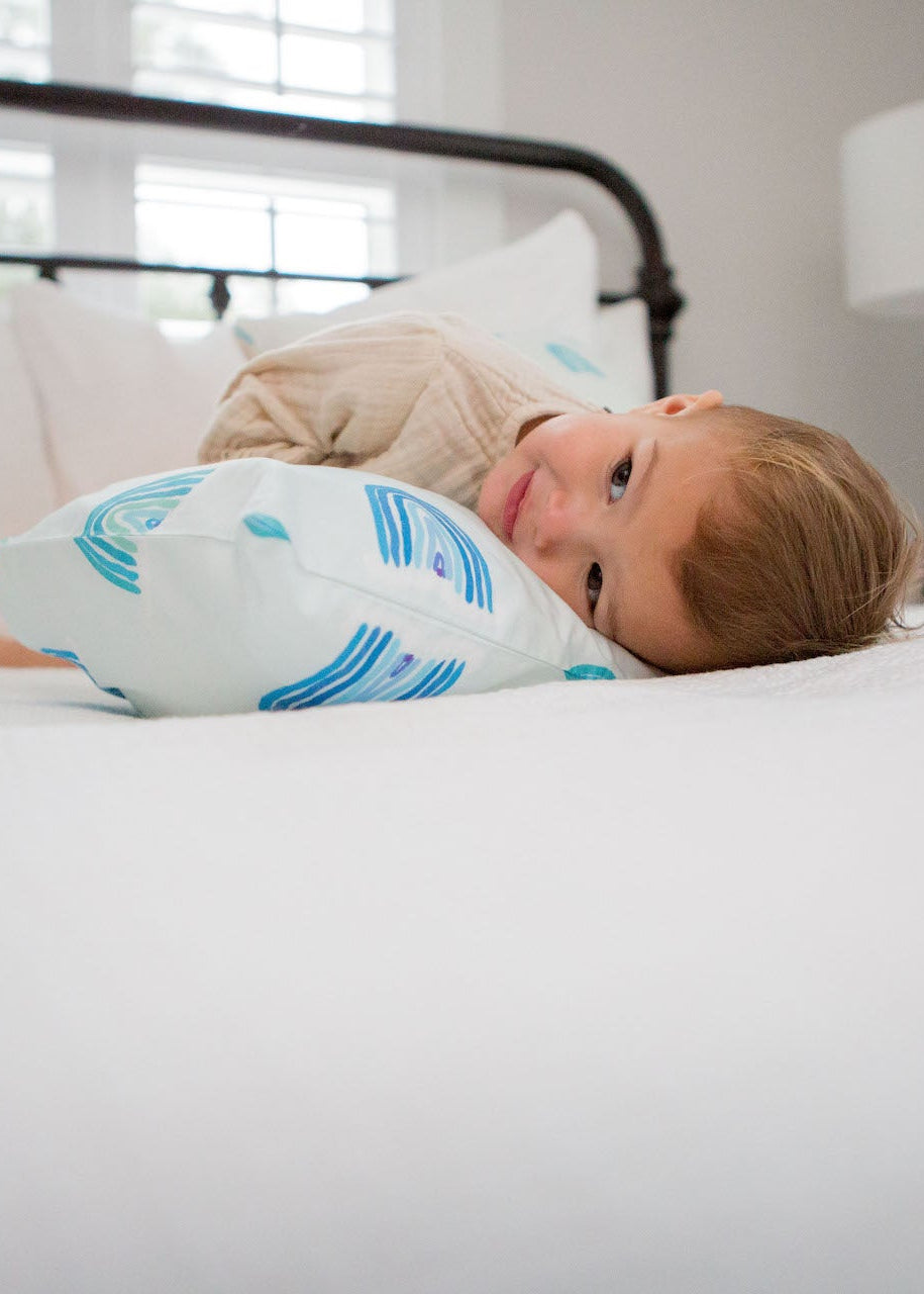 On a bed, Toddler Sleeping Pillow Cases display a front white pillow, a blue and white striped one in the middle, and three colorful ones featuring blue rainbows, yellow school buses, and pink rainbows. These organic, machine washable pillows are complemented by a thoughtful card on the bed.