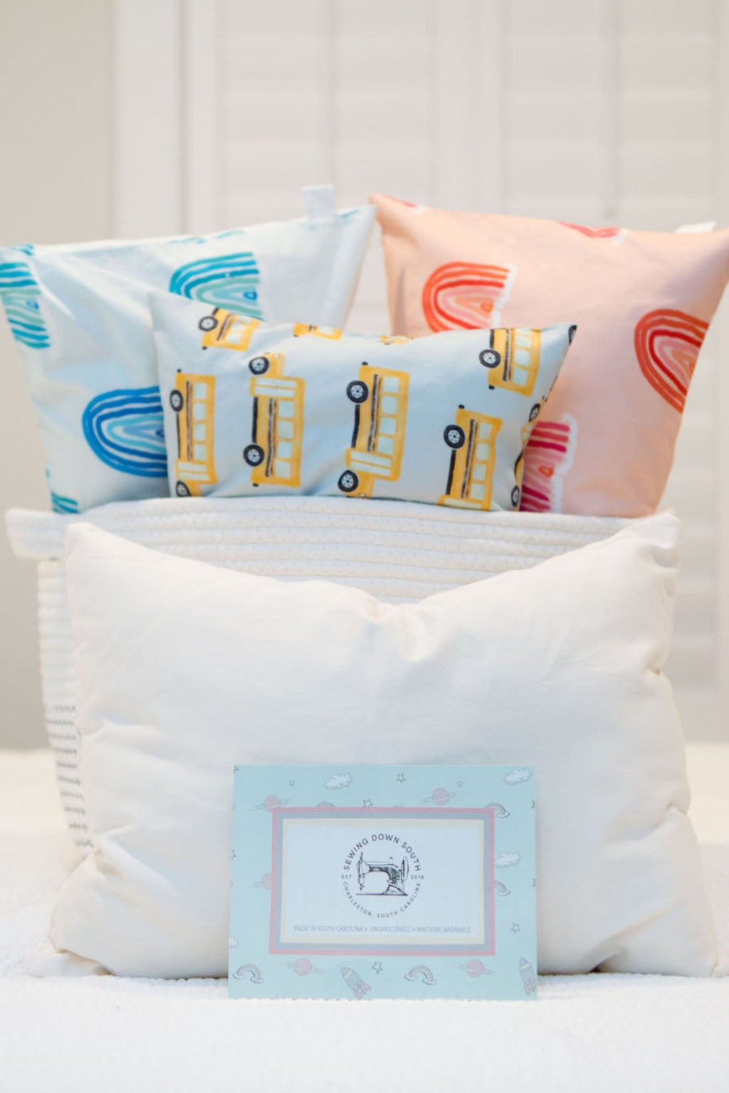 A child rests on a white bed, their head on a pillow from the Toddler Sleeping Bundle: Organic Pillow + Blue Rainbows Case. The room features muted colors, a window in the background, and a modern floor lamp beside the bed.