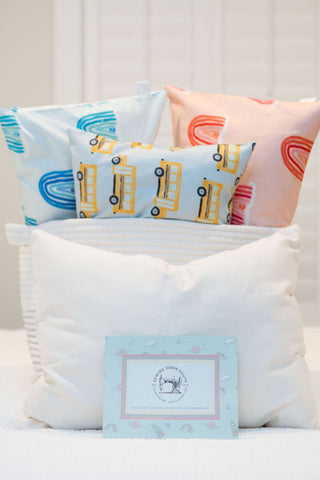 On a bed, Toddler Sleeping Pillow Cases display a front white pillow, a blue and white striped one in the middle, and three colorful ones featuring blue rainbows, yellow school buses, and pink rainbows. These organic, machine washable pillows are complemented by a thoughtful card on the bed.