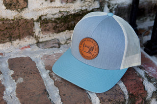 A light blue and gray trucker snapback from Sewing Down South, named Leather Patch Trucker, Grey, rests on a brick surface. The cap features a brown leather patch embossed with a fish and vintage logo text on the front.