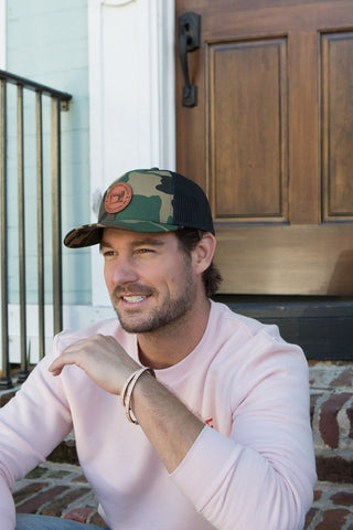 A person in a light pink sweatshirt and a Sewing Down South Leather Patch Trucker, Camo cap sits on brick steps before a wooden door, smiling and gazing into the distance with a relaxed posture. A black railing is seen on the left.