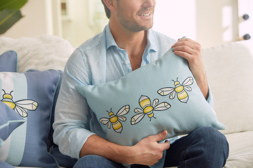 Two decorative pillows rest on a white sofa. The front pillow is a Bee Trio Lumbar Pillow from the Bee & Florals Collection, featuring a light blue surface with yellow bees. The back pillow is dark blue with light blue stripes. A window reveals part of a turquoise house.