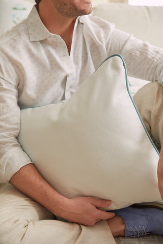A person in a light-colored, Victorian floral-patterned shirt sits on a beige couch, holding the Bee & Florals Collection: Victorian Floral in Blue Pillow with teal piping. Their head is turned away. Light brown pants and blue shoes complete the look.