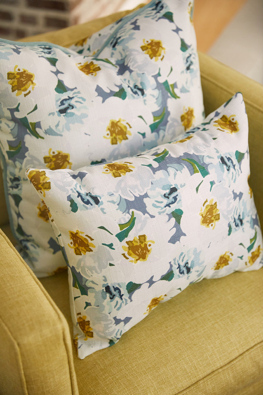 A man in a light blue shirt relaxes on a white couch, holding a floral pillow with yellow flowers. He gazes aside with a soft smile as natural light and greenery fill the room. The Bee & Florals Collection: Blossoming Peonies Lumbar Pillow beautifully complements the bright scene.