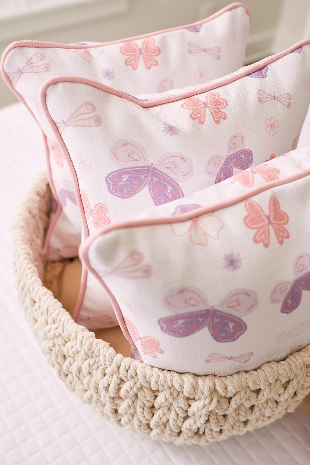 A cozy bedroom scene showcases a cute pink unicorn plush toy nestled beside two Butterfly Pillows from the Kids Collection, ideal for nursery decor. A beige knitted blanket drapes behind them, and white shutters illuminate with soft light. Perfect for any kids collection!.