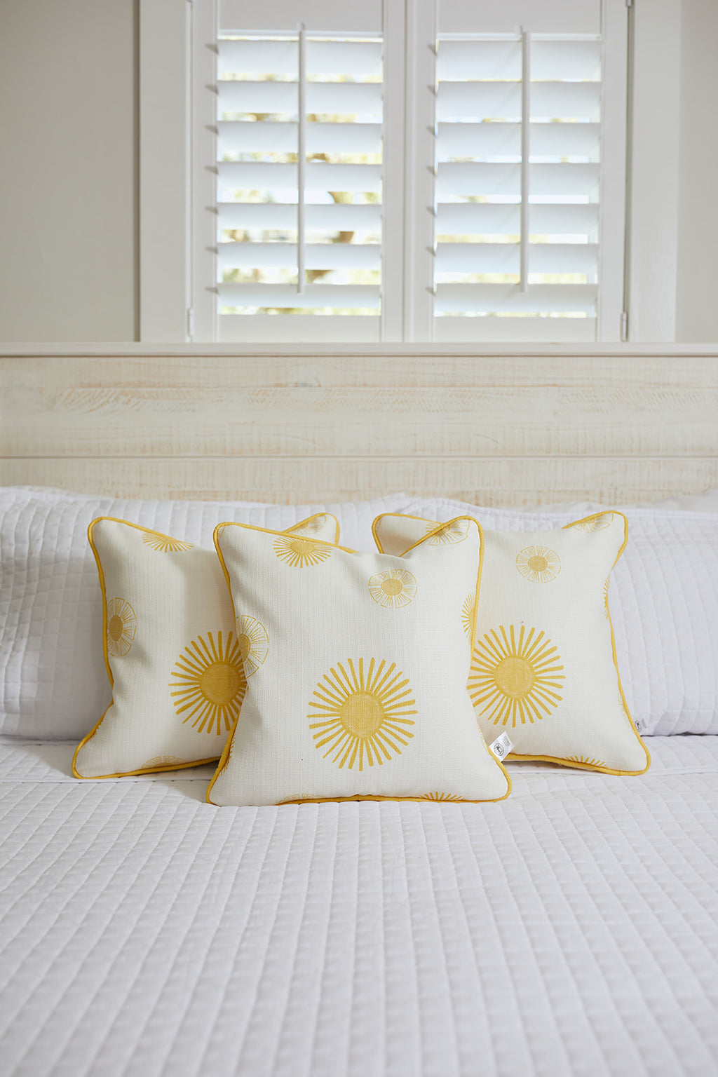 A cozy Kids Collection bedroom scene features the plush Kids Collection: Sun Pillow with sunny patterns, a fluffy bunny, and a woven basket on a white quilted bed. Sunlight streams through white shutters, creating a warm and inviting atmosphere.