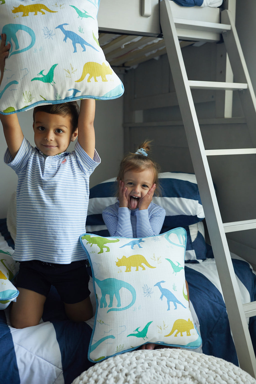 Two Kids Collection: Dinos Pillows from Sewing Down South® adorn a white bed, featuring green, blue, and yellow dinosaur silhouettes. A woven basket sits nearby, with a window framed by white shutters adding to the playful scene.