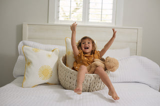 A joyful brown-haired child, clad in a yellow outfit from the Kids Collection, sits in a woven basket on a bed. Arms raised and smiling by a plush teddy bear, theyre surrounded by decorative pillows, including the Sun Pillow, all atop crisp white bedding.