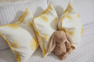 Three Sun Pillows from the Kids Collection, featuring vibrant yellow patterns, rest against a white quilt. A plush, brown bunny with floppy ears sits in front, creating a cozy and inviting atmosphere for little ones.