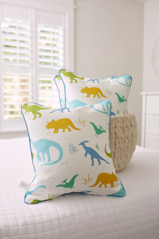 Two Kids Collection: Dinos Pillows from Sewing Down South® adorn a white bed, featuring green, blue, and yellow dinosaur silhouettes. A woven basket sits nearby, with a window framed by white shutters adding to the playful scene.