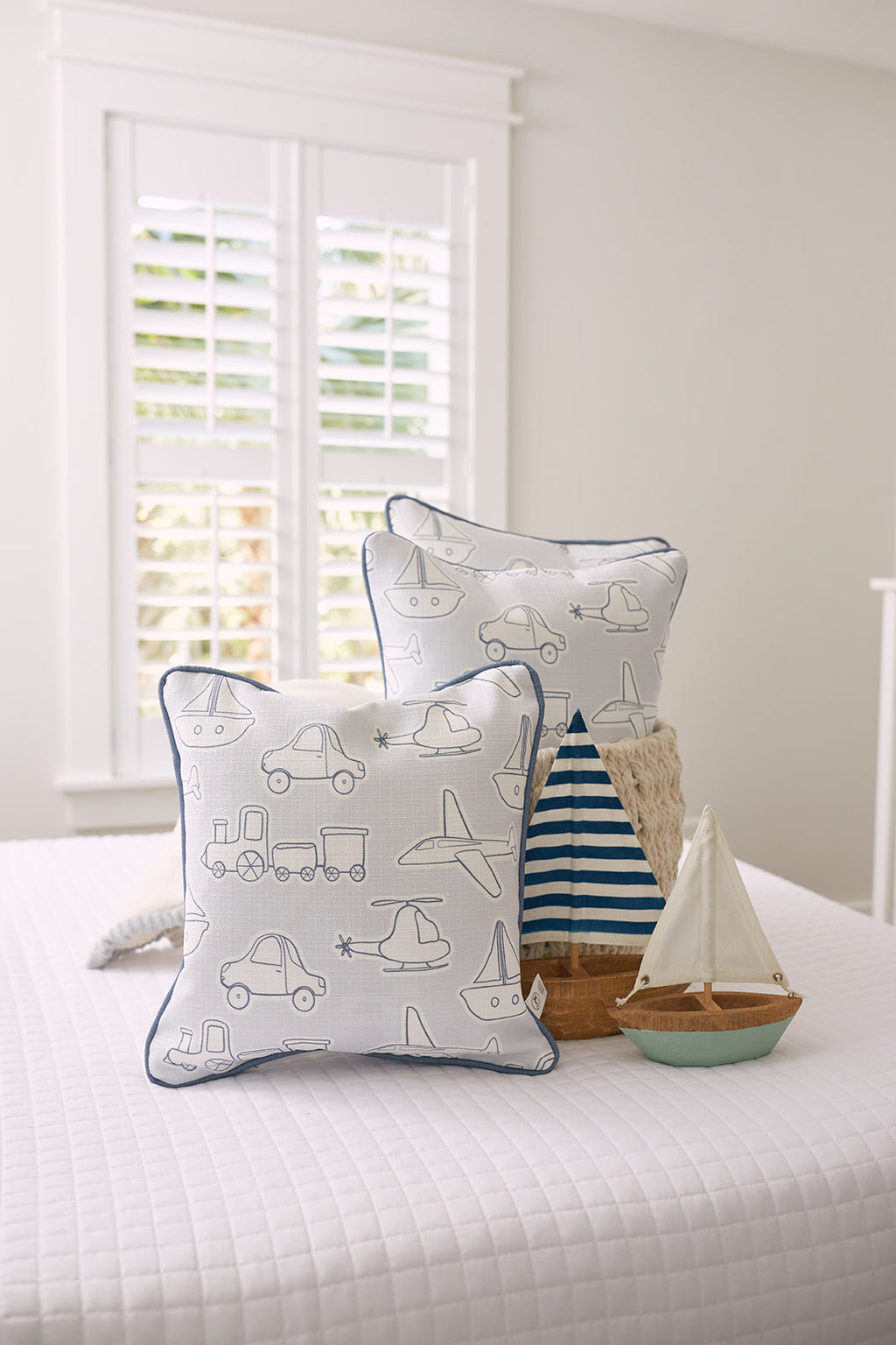 A young child with short brown hair sits on a bed, smiling widely and hugging a Kids Collection: Transportation Pillow adorned with blue and white car, train, and boat patterns that perfectly complement the striped bedding.