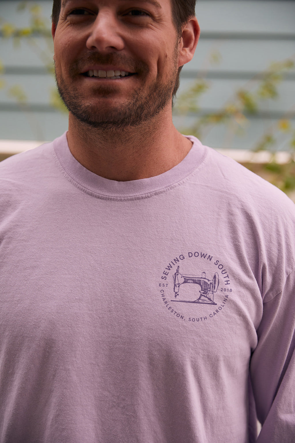 A person with short brown hair, seen from behind, wears the Whats Wrong With My Sewing? Long Sleeve Tee in Orchid. The unisex fit shirt reads the text in dark purple on 100% ring-spun cotton. The backdrop includes a blurred wall and foliage.
